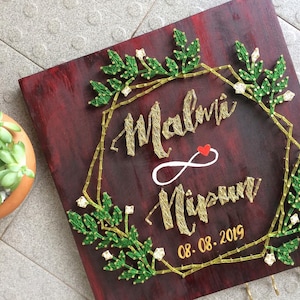Op de afbeelding: Een vierkant, donkerrood houten bord met gouden draadkunst. Het bord toont de namen "Malvi" en "Nipun", een hart en de datum "08.08.2019". Groene bladeren en een gouden geometrische rand omringen de namen. Kleine haken zijn onderaan zichtbaar.