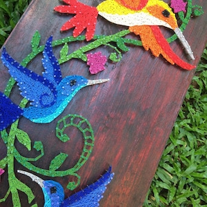 Könnte beinhalten: Ein handgefertigtes String-Art-Stück mit bunten Kolibris und floralen Elementen. Das Kunstwerk zeigt einen lebendigen Kolibri in Blau-, Rot-, Gelb- und Grüntönen mit filigranen Details und einem rustikalen Holzgrund.