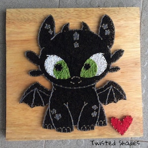 Puede incluir: Arte de hilo de un dragón negro con ojos blancos y verdes, alas y un corazón rojo sobre una tabla de madera. El dragón está hecho con hilo negro, y los ojos están detallados con hilo blanco y verde. La tabla tiene el texto "Twisted Shades".