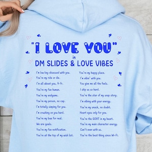 May include: A light blue hoodie with a white hood string. The back of the hoodie has the text "I LOVE YOU" in blue with hearts around it. Below the text is "in DM SLIDES & LOVE VIBES" in blue. The text continues below with a list of phrases in blue, such as "You're my ride or die" and "You're the best thing since Wi-Fi."