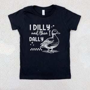 Pu&ograve; includere: T-shirt nero con la scritta bianca "I DILLY and then I DALLY", accompagnata da un'illustrazione dettagliata di un'anatra. Il design include stelle e un motivo a scacchi, creando un'estetica giocosa.