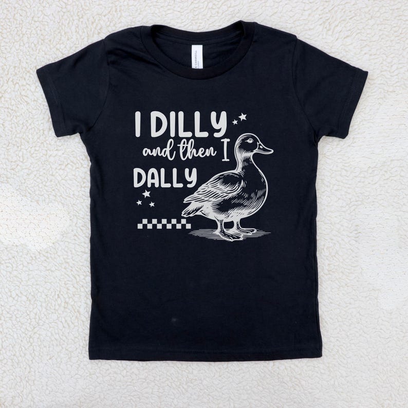 Pu&ograve; includere: T-shirt nero con la scritta bianca "I DILLY and then I DALLY", accompagnata da un'illustrazione dettagliata di un'anatra. Il design include stelle e un motivo a scacchi, creando un'estetica giocosa.