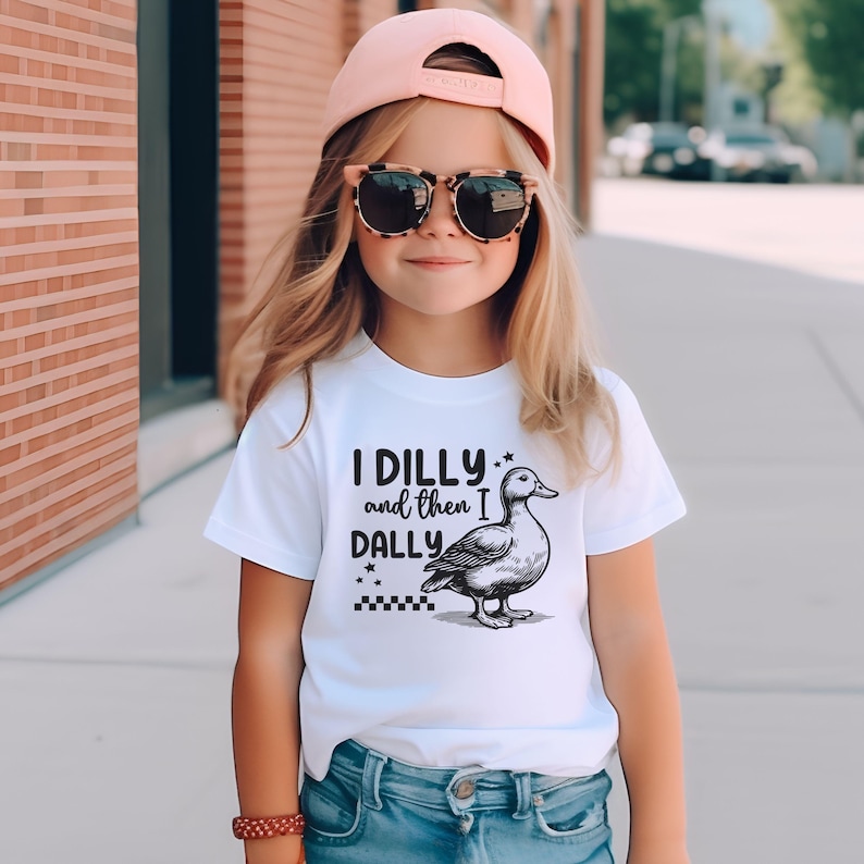 Pu&ograve; includere: Una maglietta bianca con la scritta "I DILLY and then I DALLY" e un disegno di un'anatra. Il bambino indossa un berretto da baseball rosa e occhiali da sole.