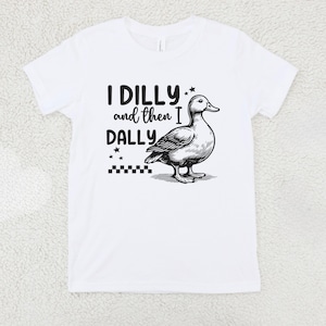 Pu&ograve; includere: T-shirt bianca con la frase "I DILLY and then I DALLY" in testo nero, accompagnata da un'illustrazione dettagliata in bianco e nero di un'anatra. Il design include stelle e un motivo a scacchiera.
