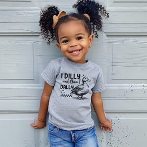 Pu&ograve; includere: Un bambino sorridente indossa una maglietta grigia con la scritta "I DILLY and then I DALLY" e un disegno di un'anatra. Il bambino indossa jeans blu e ha i capelli ricci con un fiocco.