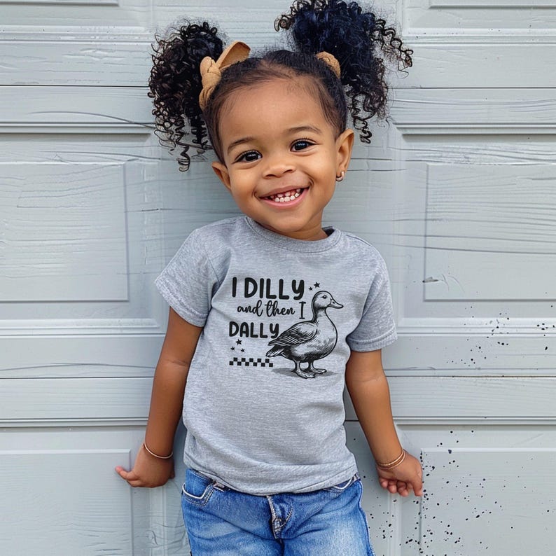 Pu&ograve; includere: Un bambino sorridente indossa una maglietta grigia con la scritta "I DILLY and then I DALLY" e un disegno di un'anatra. Il bambino indossa jeans blu e ha i capelli ricci con un fiocco.