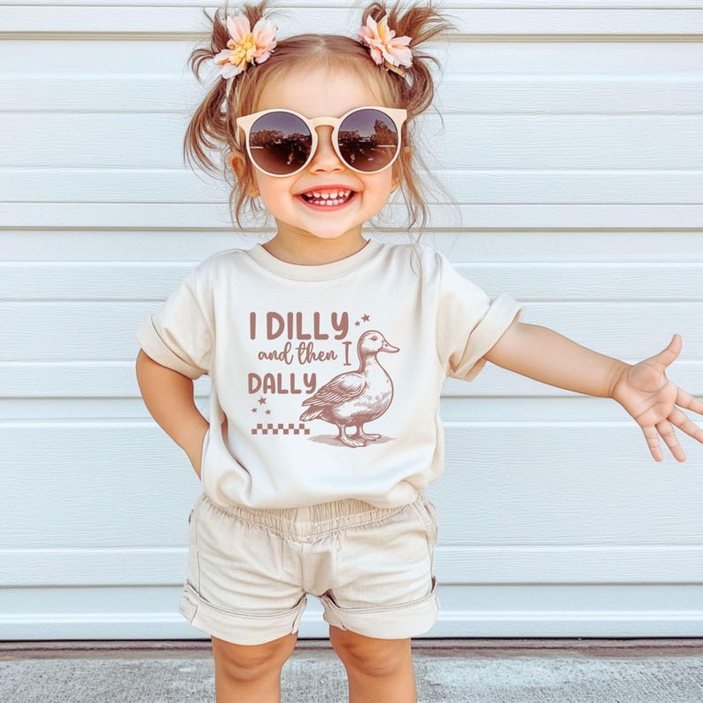Pu&ograve; includere: Un bambino piccolo indossa una maglietta e pantaloncini beige, con occhiali da sole e accessori per capelli floreali. La maglietta presenta il testo "I DILLY and then I DALLY" con un'illustrazione di anatra. Il bambino sorride e ha il braccio teso.