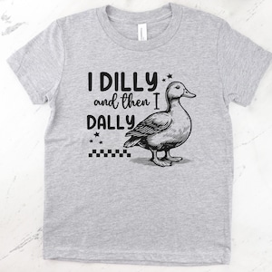 Pu&ograve; includere: T-shirt grigio chiaro con la scritta "I DILLY and then I DALLY" e un'illustrazione in bianco e nero di un'anatra. La maglietta ha un girocollo e maniche corte.