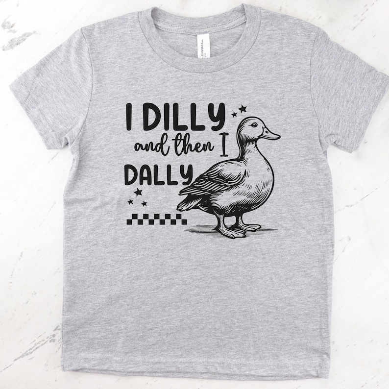Pu&ograve; includere: T-shirt grigio chiaro con la scritta "I DILLY and then I DALLY" e un'illustrazione in bianco e nero di un'anatra. La maglietta ha un girocollo e maniche corte.