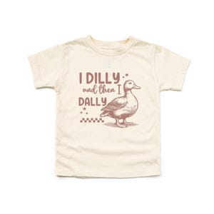 Pu&ograve; includere: T-shirt color crema per bambini piccoli con una grafica marrone di un'anatra e la scritta "I DILLY and then I DALLY". Il design include stelle e un motivo a scacchi, creando un'estetica giocosa e stravagante.