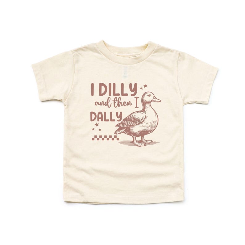 Pu&ograve; includere: T-shirt color crema per bambini piccoli con una grafica marrone di un'anatra e la scritta "I DILLY and then I DALLY". Il design include stelle e un motivo a scacchi, creando un'estetica giocosa e stravagante.