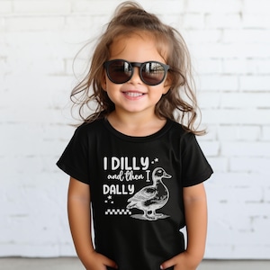 Pu&ograve; includere: T-shirt nero con la scritta "I DILLY and then I DALLY" e un'illustrazione di un'anatra. La maglietta &egrave; indossata da un bambino piccolo con occhiali da sole neri. Lo sfondo &egrave; un muro di mattoni bianchi.