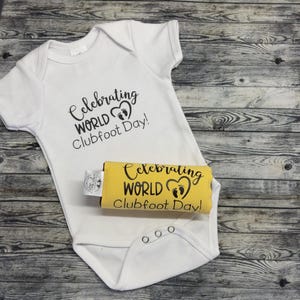 May include: A white baby onesie with black text that reads "Celebrating World Clubfoot Day!" and a black heart with footprints. A yellow fabric with the same text is in the background.
