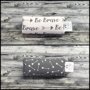 May include: Two rectangular pillows. One is white with the words "Be Brave" and arrows. The other is gray with a pattern of white arrows. Both have a small white tag with black text.