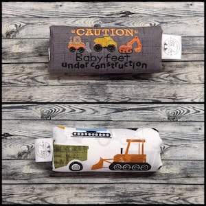 Op de afbeelding: Een rechthoekig stoffen item met twee zijden: een grijze met geborduurde bouwvoertuigen en de tekst CAUTION Baby feet under construction, en een witte met een bedrukte oranje bulldozer en groene aanhanger, beide voorzien van witte merklabeltjes.