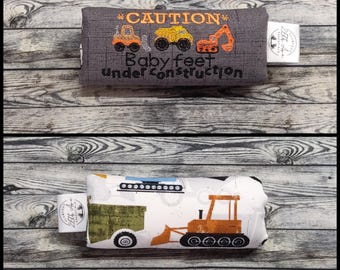 May include: Two rectangular fabric baby items. The top item is gray with embroidered construction vehicles and the text CAUTION Baby feet under construction. The bottom item is white with a printed orange bulldozer and green trailer.