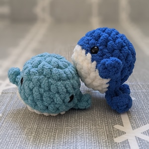 May include: Two crocheted whale plushies, one blue and one teal, with white accents and black eyes. The whales are sitting on a gray textured surface.