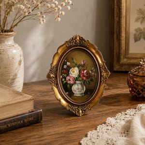 May include: An oval-shaped picture frame with an ornate, metallic gold finish and intricate scrollwork detailing. The frame contains a small, detailed oil painting depicting a bouquet of white and pink flowers arranged in a ceramic vase.