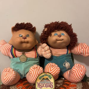 May include: Two vintage Cabbage Patch Kids Koosas dolls. One is a cat with a brown mane and the other is a lion with a brown mane. Both dolls wear striped shirts, teal corduroy overalls, and have a medallion around their necks. A Koosas label is in front.