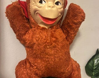 May include: Vintage plush monkey doll with a smiling face mask, red accents, and orange fur. The doll has outstretched arms and is posed against a neutral background. A classic toy.