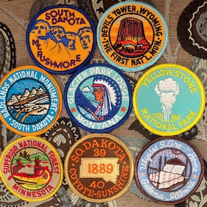 May include: Nine embroidered patches featuring various national parks and monuments. The patches are circular and feature colorful designs with text. The patches include Mount Rushmore, Devils Tower, Yellowstone National Park, Glacier Park Lodge, Badlands National Monument, Superior National Forest, and a patch with the text "S. Dakota 39 1889 40 Coyote-Sunshine".