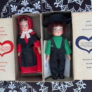 May include: Two vintage dolls dressed in traditional Amish clothing. The girl doll wears a red dress and black apron, while the boy doll wears a green shirt and black pants. Both dolls have black hats and are packaged in boxes with heart-shaped designs and text that reads "I come from Lancaster County Way Where we daily work and play My Mother early dressed me up So I could come your way".