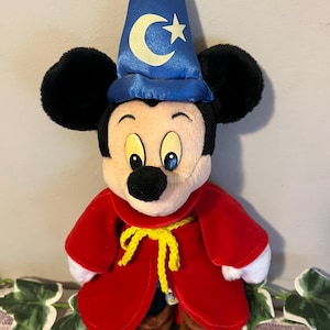 May include: A plush Mickey Mouse dressed as a wizard. He is wearing a blue hat with a crescent moon and stars, a red robe with a yellow sash, and brown shoes.