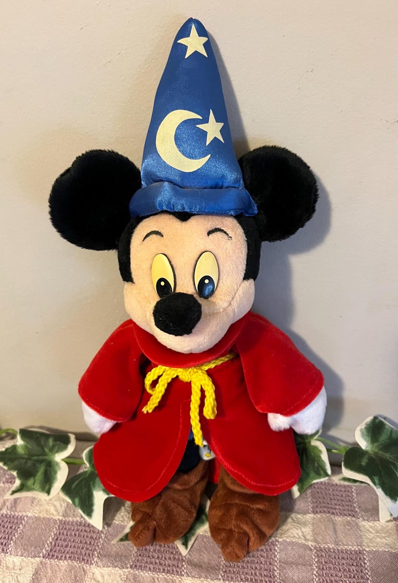 May include: A plush Mickey Mouse dressed as a wizard. He is wearing a blue hat with a crescent moon and stars, a red robe with a yellow sash, and brown shoes.