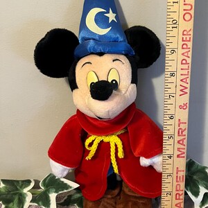 May include: A plush Mickey Mouse dressed as a wizard. He is wearing a blue hat with a crescent moon and stars, a red robe with a yellow sash, and brown shoes. He is approximately 10 inches tall.