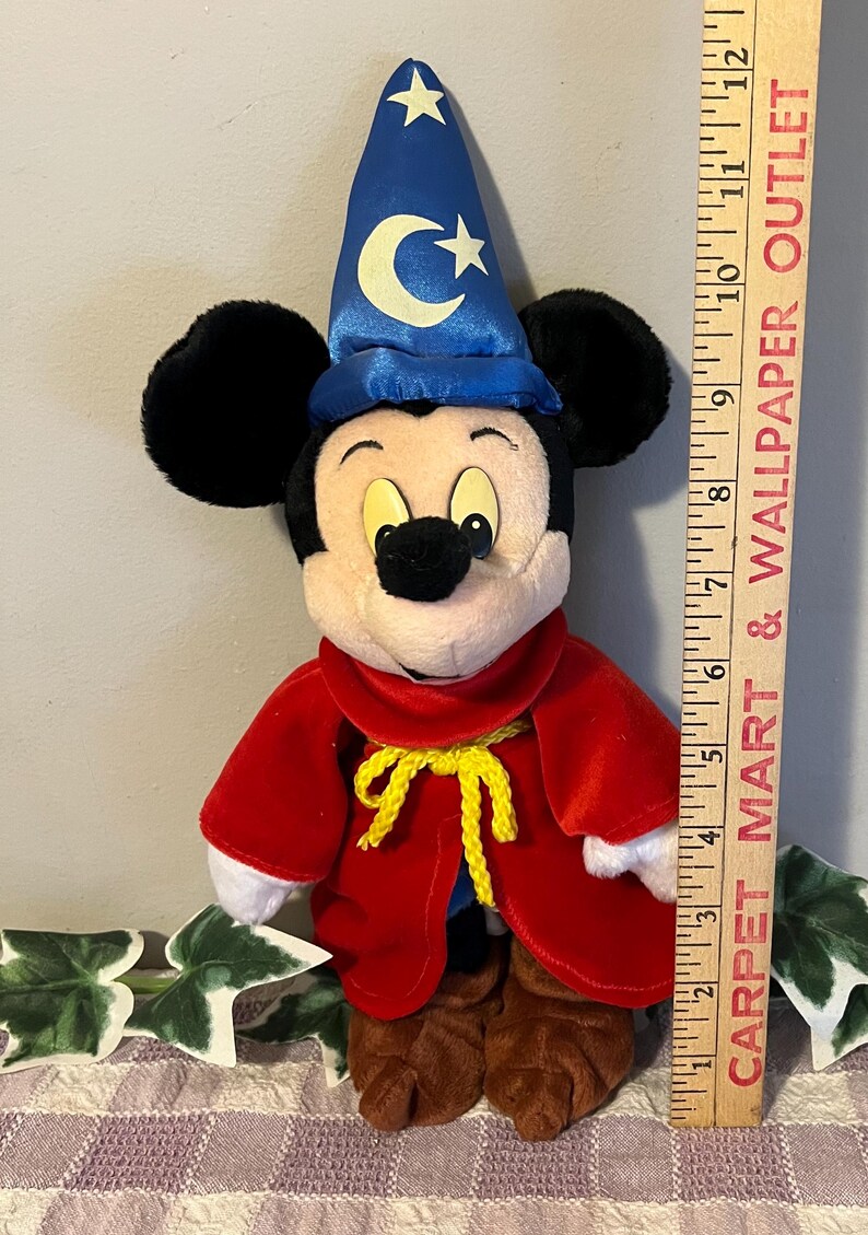 May include: A plush Mickey Mouse dressed as a wizard. He is wearing a blue hat with a crescent moon and stars, a red robe with a yellow sash, and brown shoes. He is approximately 10 inches tall.
