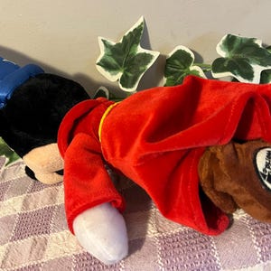 May include: A plush Mickey Mouse toy wearing a blue wizard hat and a red robe. The toy has a brown nose and black eyes. The robe has a gold trim. The toy is lying on a purple and white checkered fabric. The toy has a tag that says "Disney World"