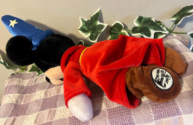 May include: A plush Mickey Mouse toy wearing a blue wizard hat and a red robe. The toy has a brown nose and black eyes. The robe has a gold trim. The toy is lying on a purple and white checkered fabric. The toy has a tag that says "Disney World"