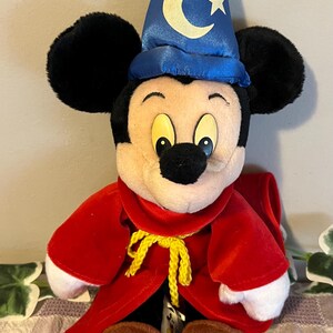 May include: A plush Mickey Mouse dressed as a wizard. He is wearing a blue hat with a crescent moon and stars, a red robe with a yellow rope tie, and brown boots.