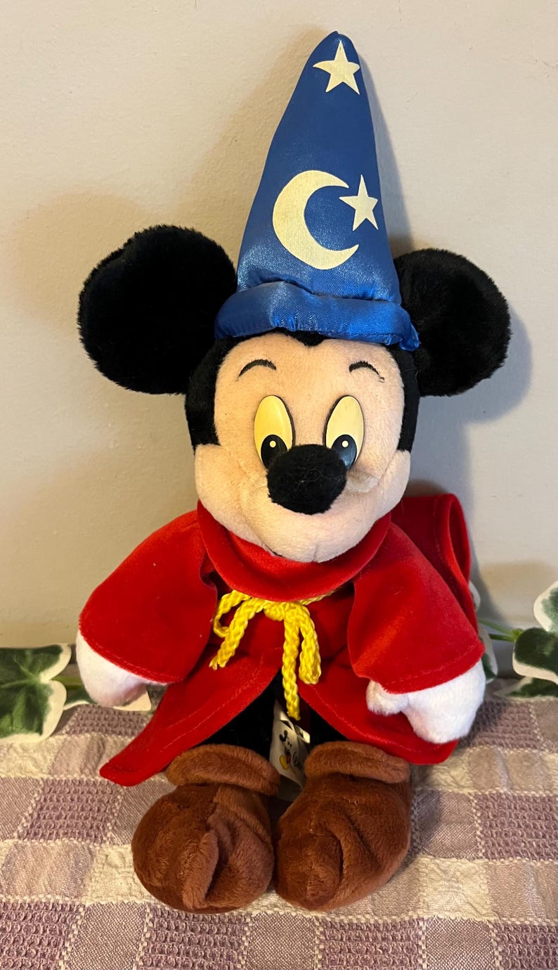May include: A plush Mickey Mouse dressed as a wizard. He is wearing a blue hat with a crescent moon and stars, a red robe with a yellow rope tie, and brown boots.