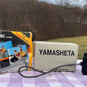 May include: A black and silver 35mm film camera with a black strap. The camera is sitting on a white and purple checkered surface. The camera has the words "PENTAX" and "Auto-Focus 35" printed on it. The camera is next to a white box with the word "YAMASHETA" printed on it. The box is next to a yellow box with the words "Kodak tele disc camera" printed on it.