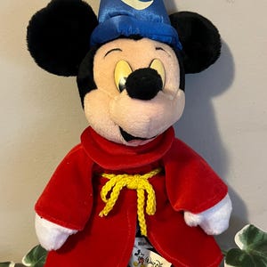 May include: A plush Mickey Mouse dressed as a wizard. He is wearing a blue hat with stars and a crescent moon, a red robe with a yellow rope tie, and white gloves. The plush toy has a "Walt Disney World" tag attached to its robe.