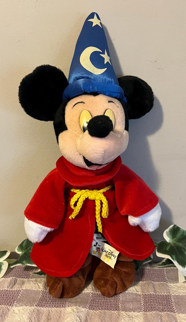 May include: A plush Mickey Mouse dressed as a wizard. He is wearing a blue hat with stars and a crescent moon, a red robe with a yellow rope tie, and white gloves. The plush toy has a "Walt Disney World" tag attached to its robe.