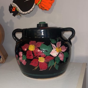 May include: Black ceramic crock with a lid, hand-painted with pink, red, and white flowers with yellow centers and green leaves.