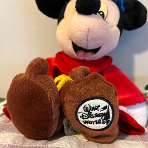 May include: A plush Mickey Mouse wearing a red robe and a blue hat. He is sitting with his legs crossed and has a brown patch on his feet that says "Walt Disney World".