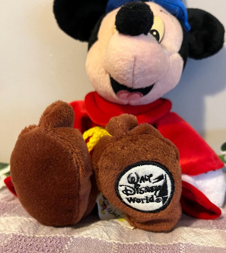 May include: A plush Mickey Mouse wearing a red robe and a blue hat. He is sitting with his legs crossed and has a brown patch on his feet that says "Walt Disney World".