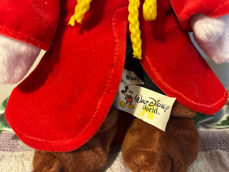 May include: Red plush Mickey Mouse wearing a red robe with yellow trim and a black hat. The plush toy has a "Mickey's" tag and a "Walt Disney World" tag attached to the robe.