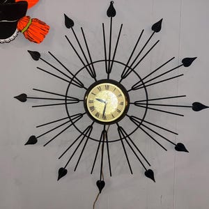 May include: A black metal starburst clock with a gold-toned face and black hands. The clock has a circular design with radiating lines ending in heart-shaped points. A vintage Halloween decoration is visible in the upper left corner.