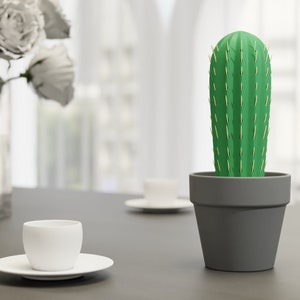3D-gedruckter Kaktus Zahnstocherhalter in Topffarbe Grau auf einem Tisch neben einer weißen Tasse, imitiert mit herauskommenden Zahnstochern eine Kaktuspflanze.