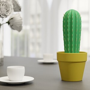 3D-gedruckter Kaktus Zahnstocherhalter in Topffarbe Gelb auf einem Tisch neben einer weißen Tasse, imitiert mit herauskommenden Zahnstochern eine Kaktuspflanze.