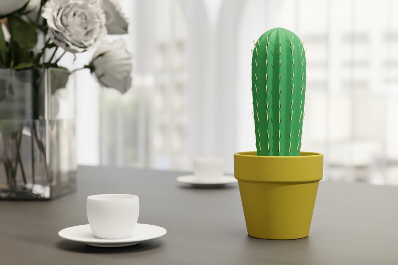 3D-gedruckter Kaktus Zahnstocherhalter in Topffarbe Gelb auf einem Tisch neben einer weißen Tasse, imitiert mit herauskommenden Zahnstochern eine Kaktuspflanze.