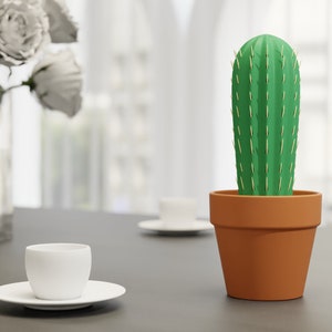 3D-gedruckter Kaktus Zahnstocherhalter in Topffarbe Orange auf einem Tisch neben einer weißen Tasse, imitiert mit herauskommenden Zahnstochern eine Kaktuspflanze.