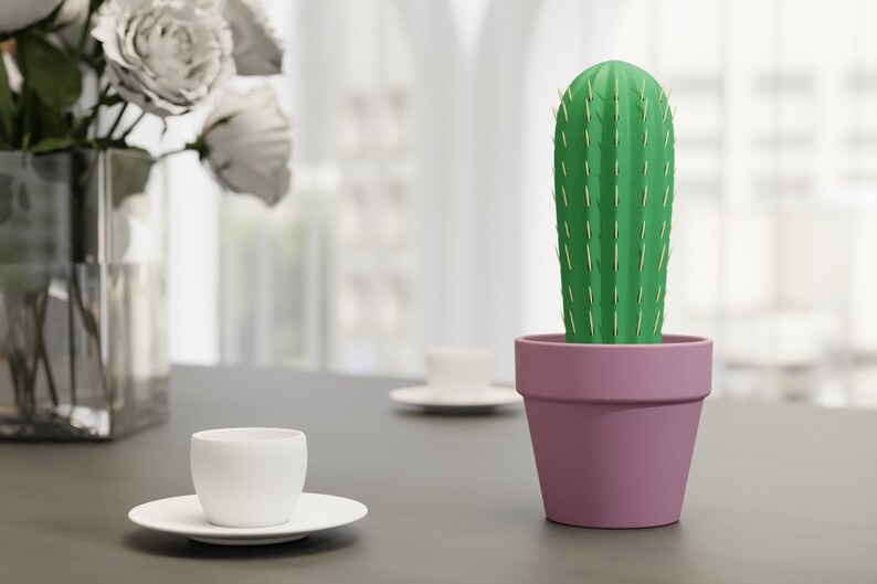 3D-gedruckter Kaktus Zahnstocherhalter in Topffarbe Pink auf einem Tisch neben einer weißen Tasse, imitiert mit herauskommenden Zahnstochern eine Kaktuspflanze.