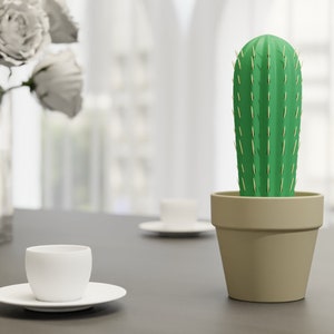 3D-gedruckter Kaktus Zahnstocherhalter in Topffarbe Sand auf einem Tisch neben einer weißen Tasse, imitiert mit herauskommenden Zahnstochern eine Kaktuspflanze.