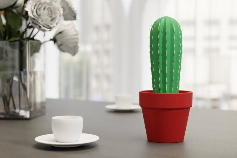 3D-gedruckter Kaktus Zahnstocherhalter in Topffarbe Rot auf einem Tisch neben einer weißen Tasse, imitiert mit herauskommenden Zahnstochern eine Kaktuspflanze.