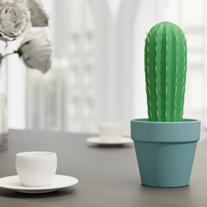 3D-gedruckter Kaktus Zahnstocherhalter in Topffarbe Eisblau auf einem Tisch neben einer weißen Tasse, imitiert mit herauskommenden Zahnstochern eine Kaktuspflanze.